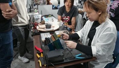 Gelissa Loh in a white shirt solders wires at a workbench, with other students observing in the background.