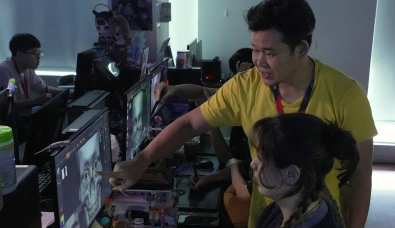 Julian Khor in a yellow shirt points to a 3D model of a skull on a student's computer screen in a classroom setting.