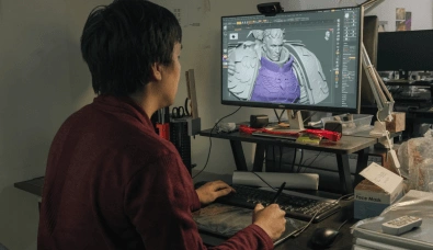 Teo Chia Kai in a red shirt works on a 3D model of a character with armor on a computer screen, using a graphics tablet.
