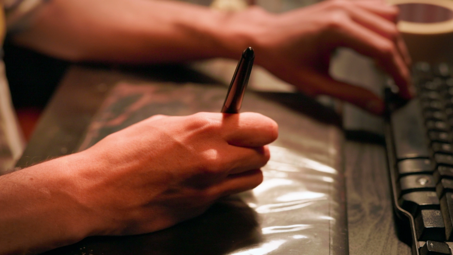 A close-up of a hand holding a pen, poised over a glossy surface, while another hand types on a computer keyboard.