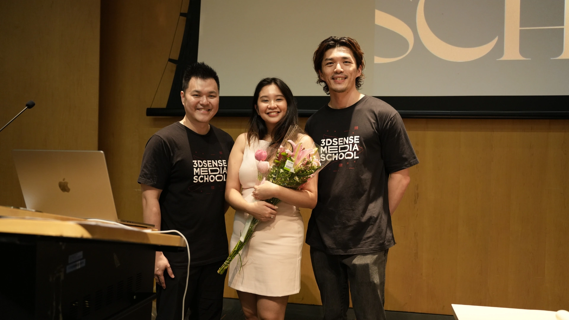 Student stands between two 3dsense lecturers on stage during graduation, holding a bouquet and smiling towards the camera.
