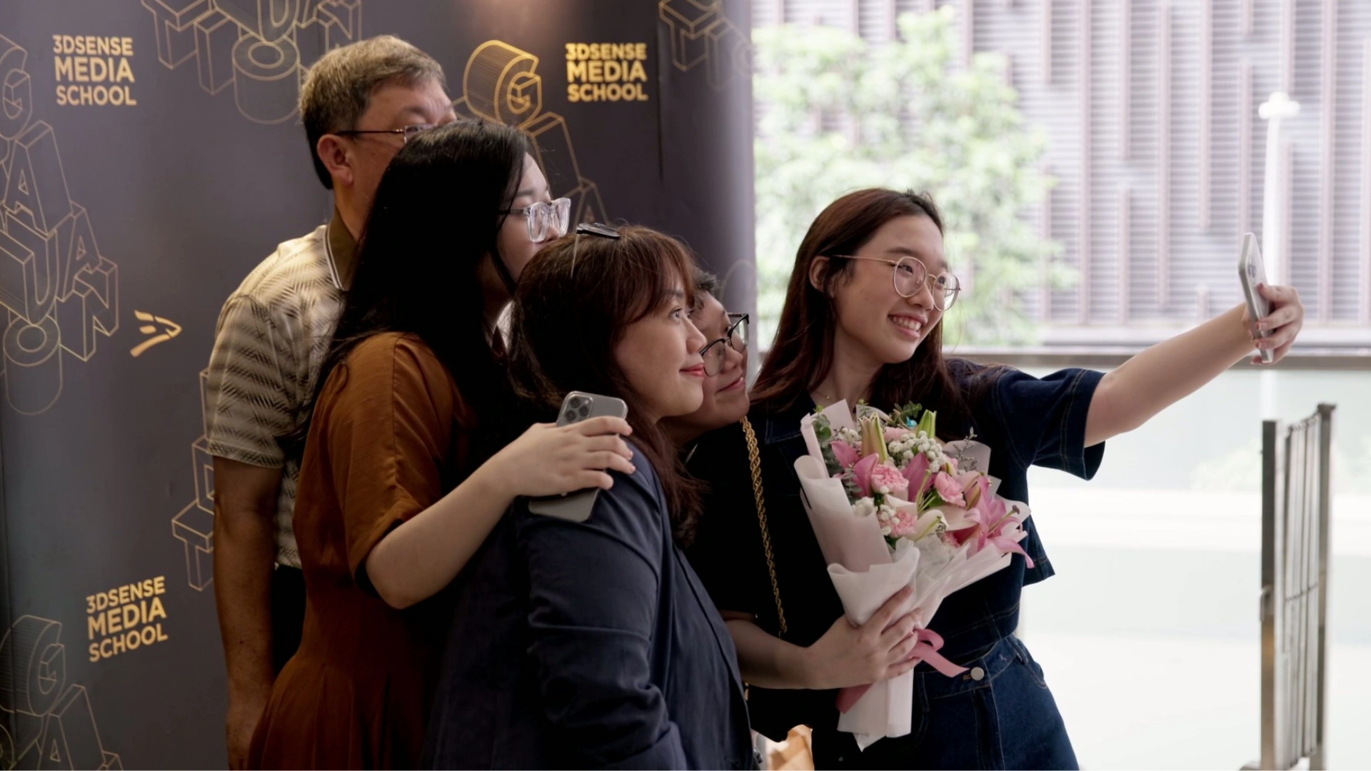 A graduate and her family pose for a selfie at the 3dsense graduation, holding a bouquet in front of a branded backdrop.