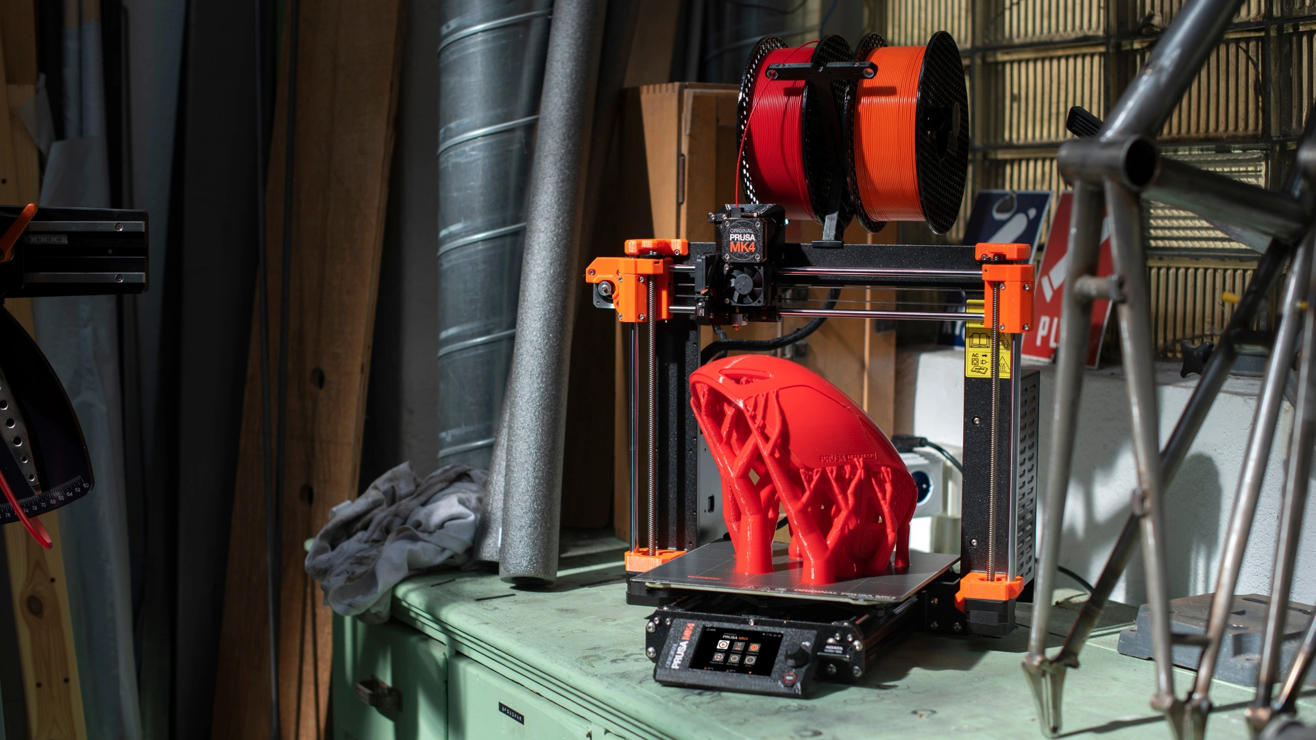 A 3D printer with vibrant orange accents produces a detailed red object, surrounded by tools and materials in a workshop.