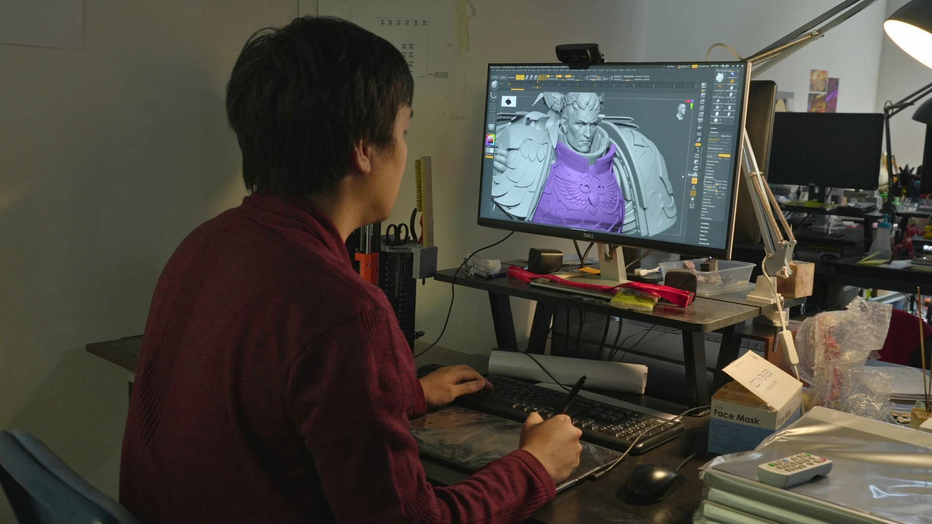 A lecturer working at a desk, using a tablet to design a 3D model of a detailed armored figure on a computer screen.