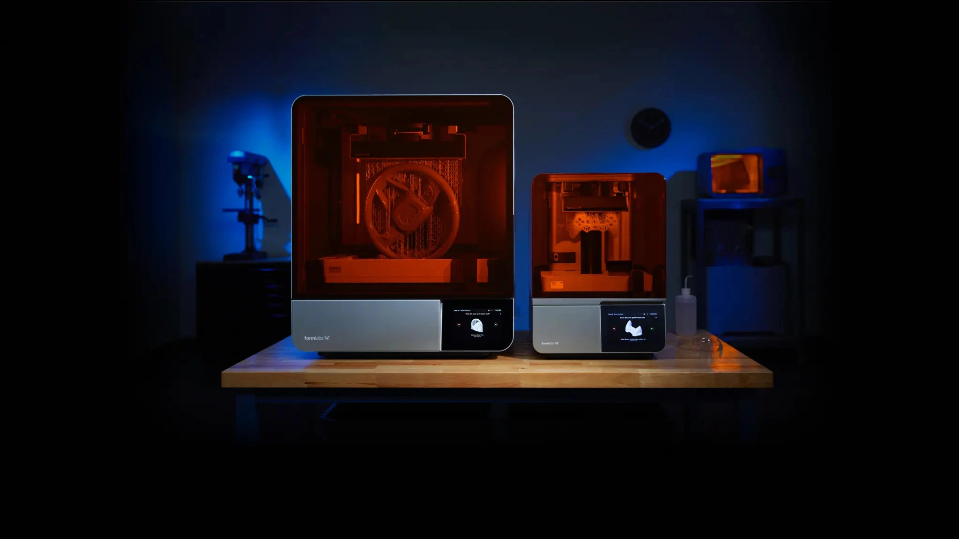 Two illuminated 3D printers sit on a wooden table, showcasing intricate models against a backdrop of blue lighting.