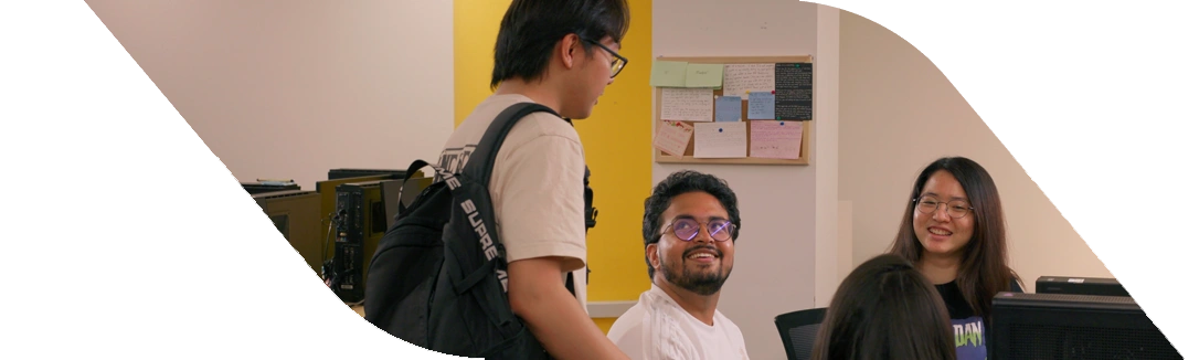 A group of students engaged in discussion near computers, with a yellow wall and a bulletin board in the background.