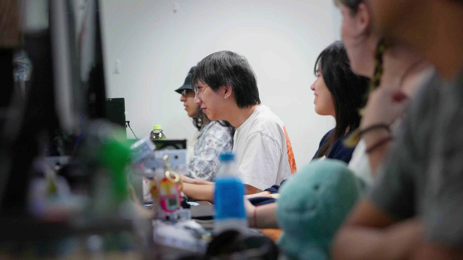 A group of students is seated at desks, focused on their screens, with various beverages and a plush toy visible in the foreground.