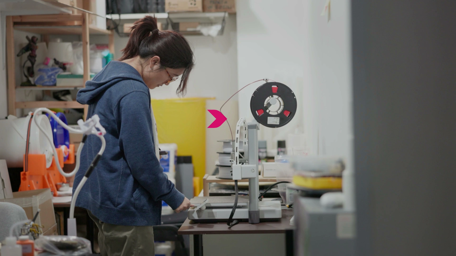 A person works at a table with a 3D printer and filament spool, surrounded by tools and supplies in a workshop setting.