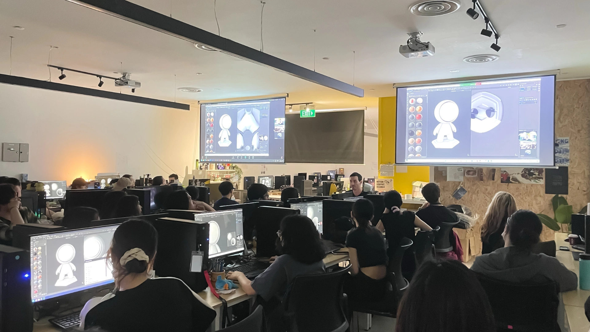 A classroom filled with students focused on computer screens displaying 3D modeling software, with a large projector showing designs.