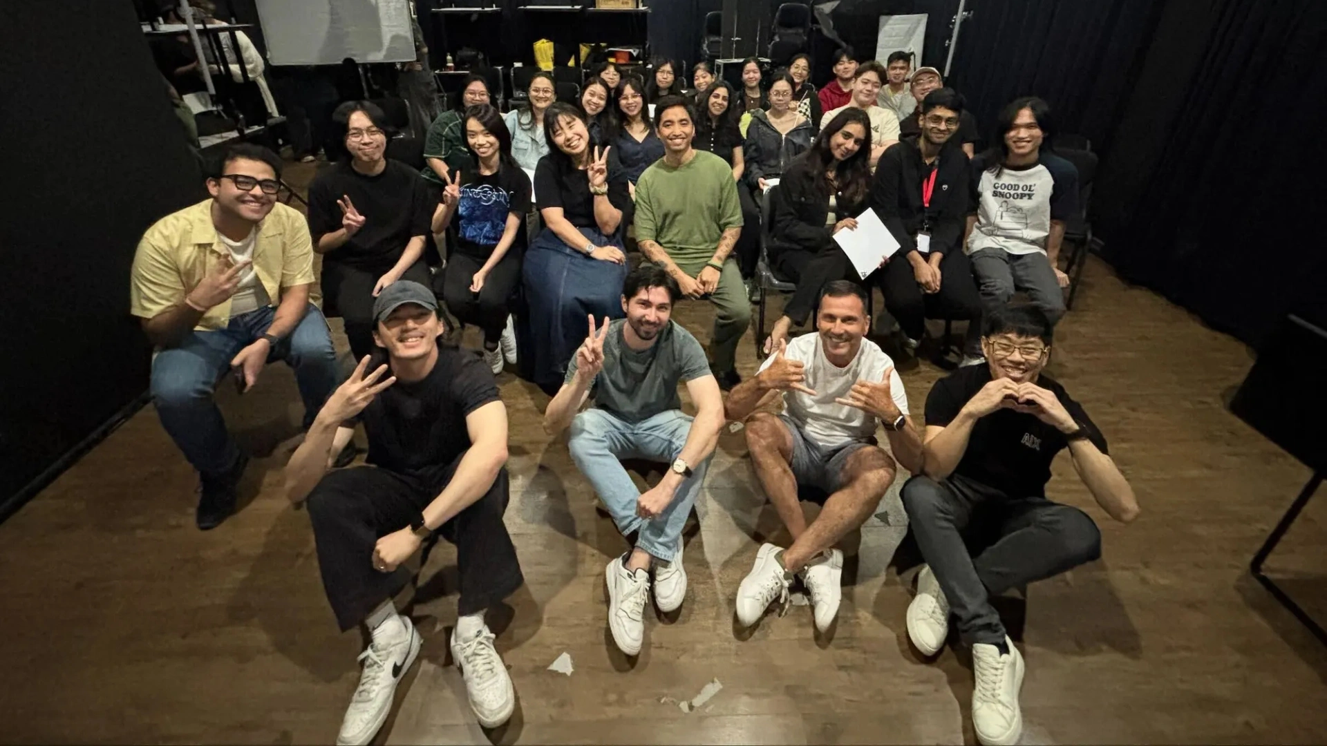 A diverse group of individuals sit and pose together in a theater-like setting, showing peace signs and playful gestures.