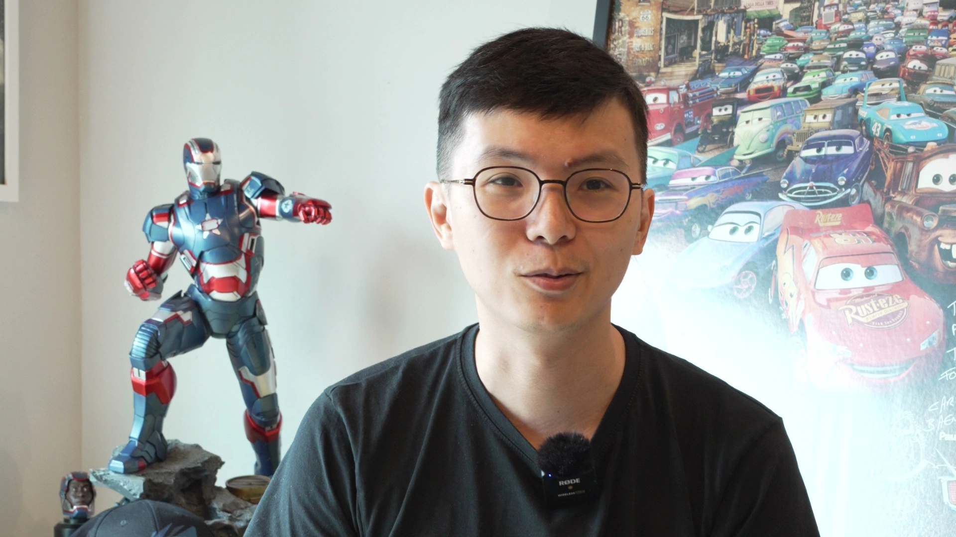 A person wearing a black shirt sits in front of a colorful Cars movie poster, with an Iron Man figurine displayed beside them.