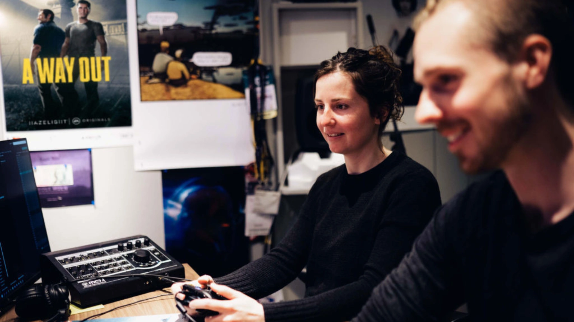 A person plays a video game with a controller, while another sits nearby, surrounded by gaming posters and equipment.