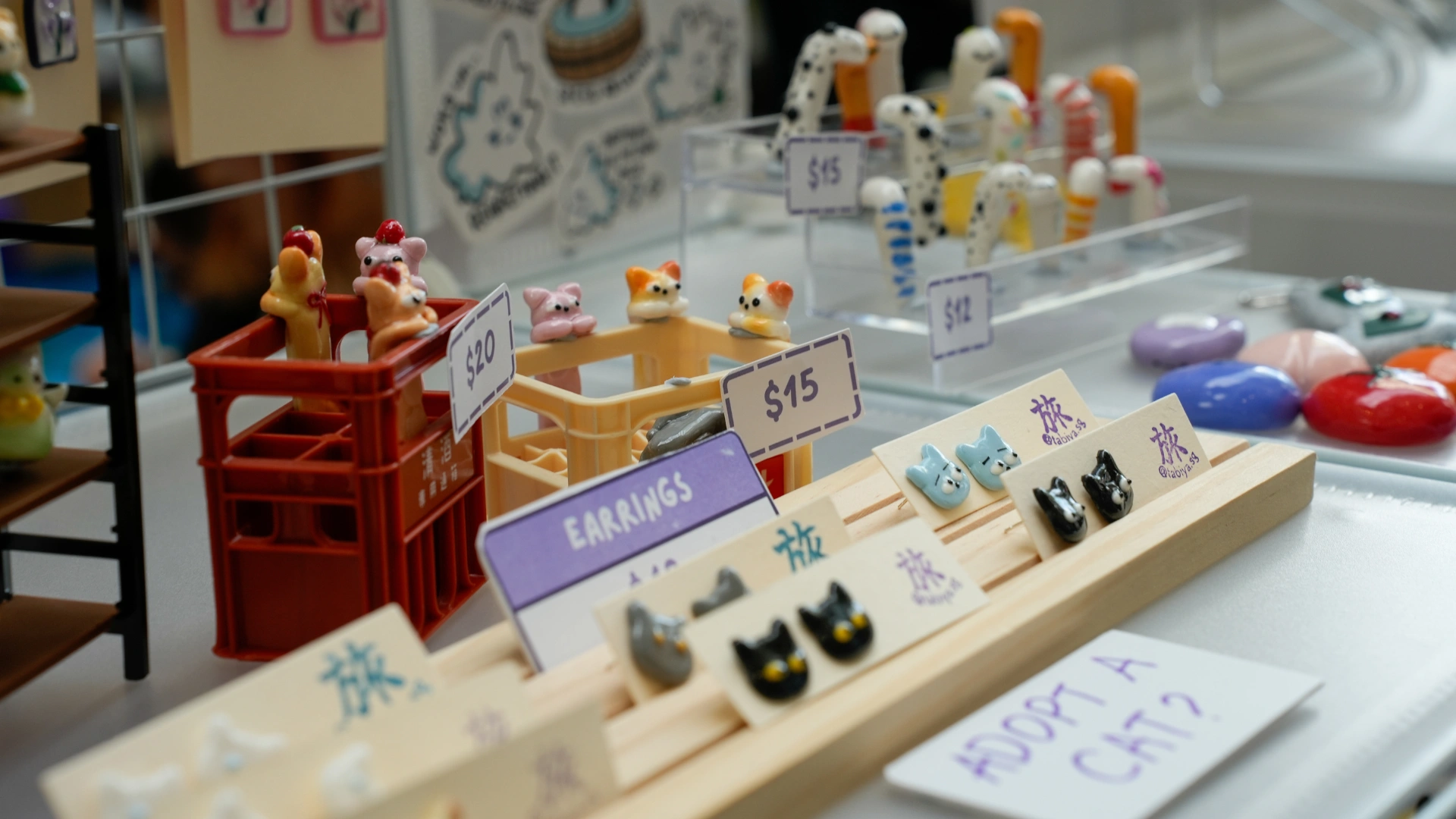 A colorful display of handmade animal-themed earrings and figurines, with various price tags, inviting visitors to adopt a cat.