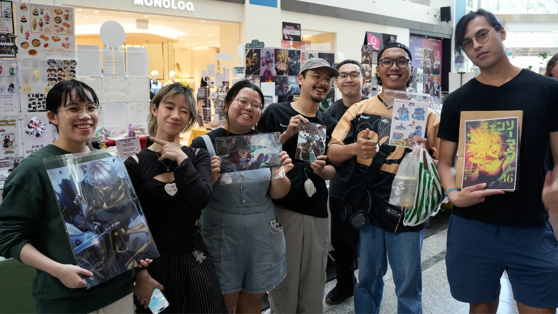 A group of fans proudly display anime merchandise including posters and stickers at a vibrant event in a mall.