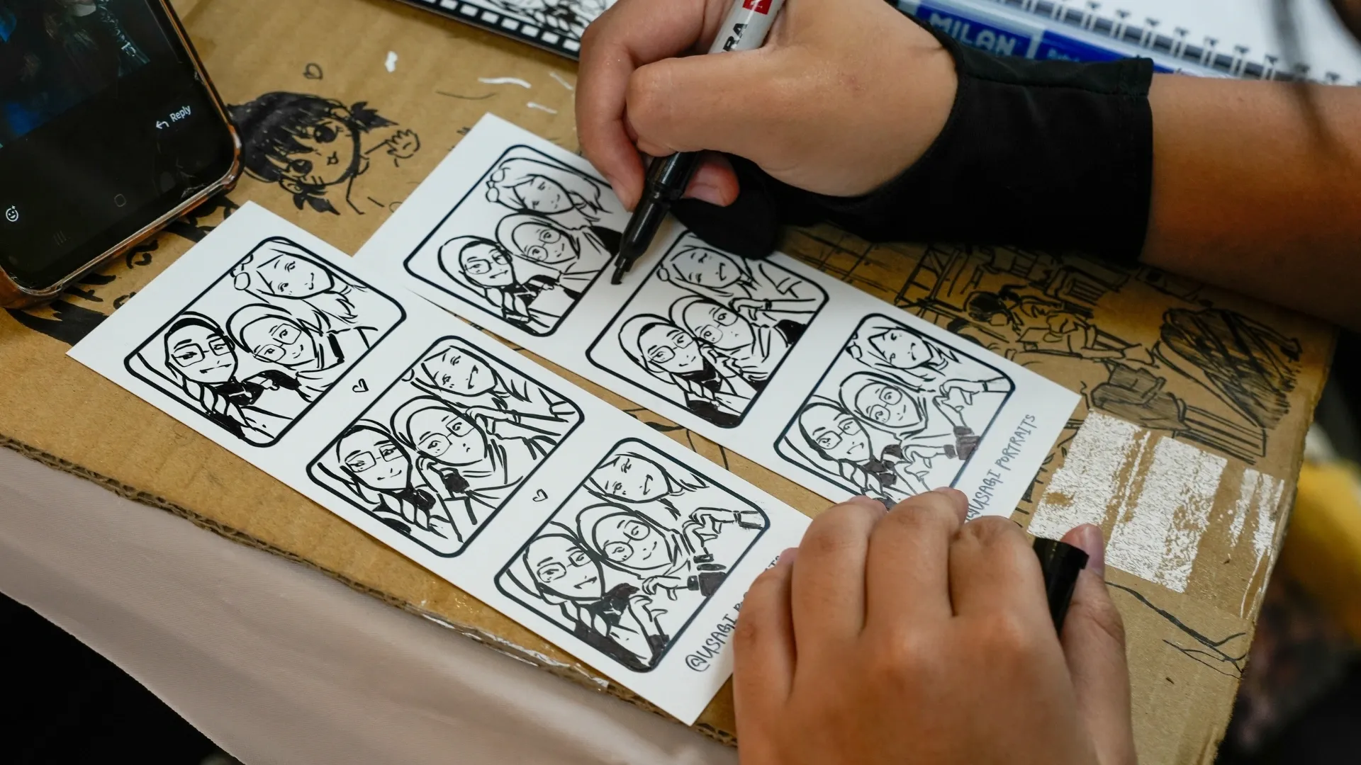 A person sketches cartoon-style self-portraits on stickers while holding a marker, with a phone and sketchpad nearby.