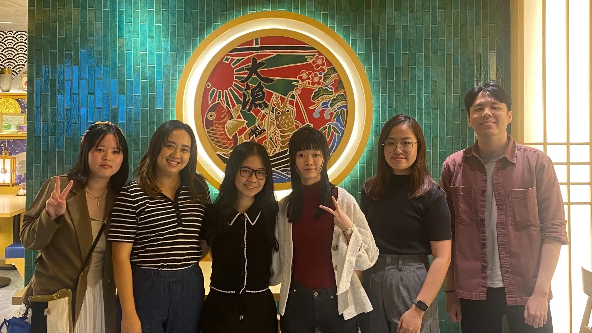 A group of six 3dsense alumni stands in front of a colorful mural featuring fish and floral designs, showcasing vibrant decor in a relaxed atmosphere.