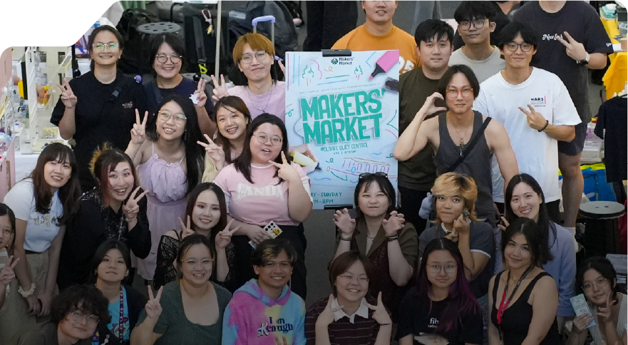 A diverse group of people poses together at a 3dsense Makers Market, smiling and making peace signs, surrounded by various booths and stalls.